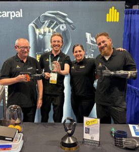 Four Metacarpal team members stood behind their table at a trade show, holding their winning trophy for the GEM hand.